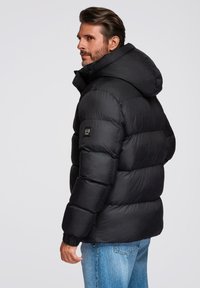Chaquetón negro acolchado con capucha, que presenta secciones acolchadas, puños de canalé y un pequeño parche con el logo en la manga izquierda.