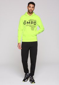 Felpa verde neon con testo grafico nero, abbinata a pantaloni tuta neri e scarpe da ginnastica nere con accenti neon. Completo di abbigliamento attivo casual.