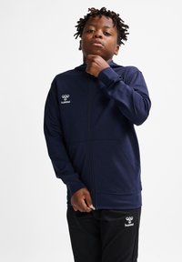 Dunkelblaues Zip-Hoodie aus glattem Stoff, mit Kängurutaschen und Logo auf der Brust. Getragen mit schwarzen Hosen und Händen, die den Reißverschluss anpassen.