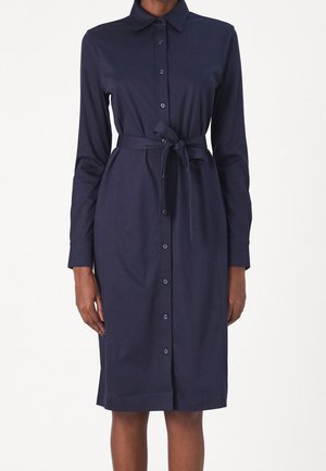 Femme portant une robe chemise bleu marine mi-longue, à manches longues, avec une ceinture à nouer et un col pointu.