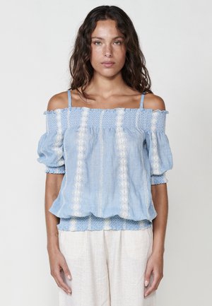 Blouse bleu clair décolletée avec des manches bouffantes, ornée de motifs brodés blancs et d'une finition texturée. Tissu doux et coupe décontractée.