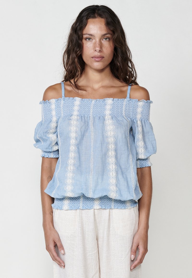 Blusa azzurro chiaro fuori spalla con maniche a sbuffo, caratterizzata da motivi ricamati bianchi e una finitura testurizzata. Tessuto morbido e vestibilità comoda.