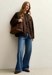 Chaqueta marrón texturizada sobre un top marrón, combinada con jeans azul acampanados y zapatos marrones oscuros. Sosteniendo un bolso tote marrón. Fondo claro.