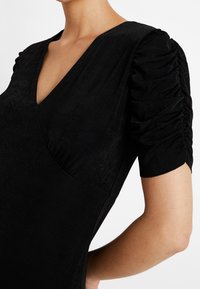 Sort kjole med V-udskæring, korte pufærmer med indsamlet detaljer og en glat tekstur. Den tætsiddende design fremhæver silhouette.