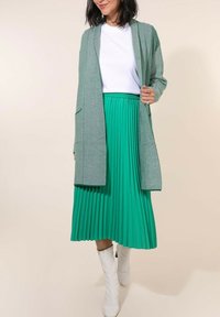 Jupe plissée verte avec une taille haute, associée à un cardigan rayé gris. La tenue comprend des bottes blanches jusqu'aux genoux et un haut blanc.