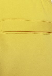 Tela de algodón amarilla con una textura suave, que presenta un bolsillo horizontal y un detalle de dobladillo cosido. Color uniforme sin patrones visibles.