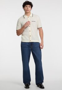 Cremefarbenes Kurzarm-Shirt mit Knopfleiste und schwarzem Besatz sowie dem Aufdruck "GUESS JEANS", kombiniert mit dunkelblauen, weit geschnittenen Jeans und schwarzen Schuhen.