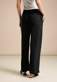 Schwarze weit geschnittene Hose mit strukturiertem Zickzackmuster, ausgestattet mit Seitentaschen und hohem Bund. Tragen mit flachen Sandalen.
