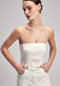 Top senza spalline bianco con cuciture verticali, caratterizzato da accenti in metallo dorato e una texture liscia, completato da una vestibilità sartoriale.