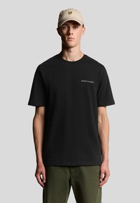 Camiseta negra de cuello redondo con mangas cortas y logo blanco "LYLE & SCOTT", combinada con pantalones verde oliva y una gorra de color beige claro con un logo de mariposa.