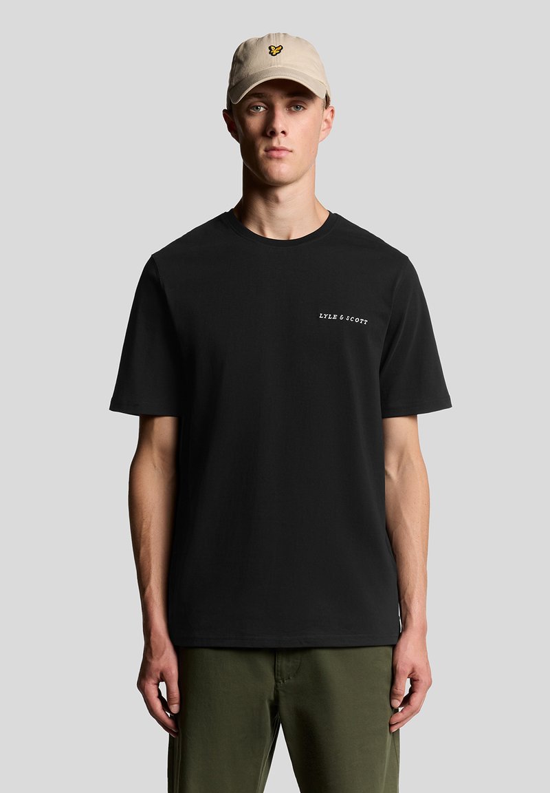 Camiseta negra de cuello redondo con mangas cortas y logo blanco "LYLE & SCOTT", combinada con pantalones verde oliva y una gorra de color beige claro con un logo de mariposa.