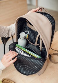 Beige backpack with a smooth exterior. Inside features a black lining, water bottle, notebooks, and a zippered pocket for accessories.