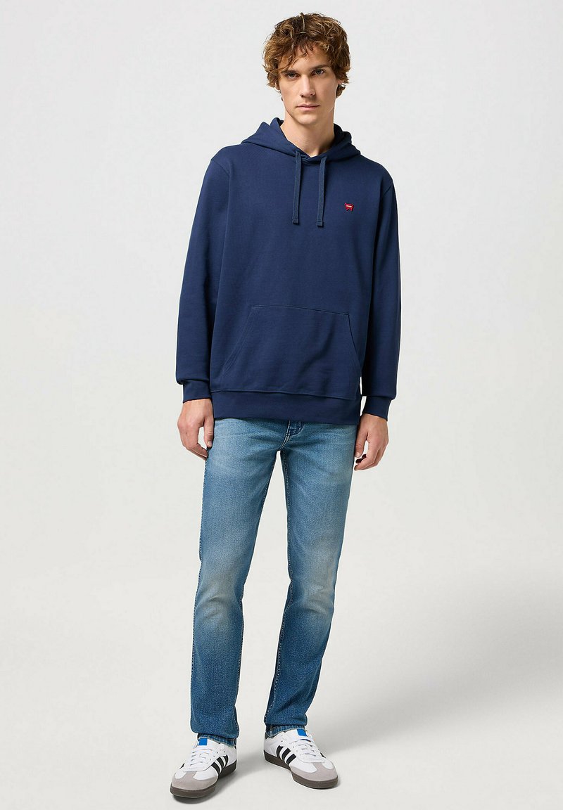 Granatowy hoodie z miękkiego materiału, z kieszenią z przodu, sznurków oraz małym logo na klatce piersiowej, w połączeniu z jasnoniebieskimi dżinsami i białymi sneakersami.