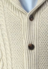 Gilet en tricot beige avec motif de câble et texture côtelée, doté de deux boutons foncés à l'avant et d'un col châle.