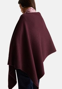 Donna che indossa un poncho borgogna sopra una sciarpa rosa e pantaloni scuri, vista di spalle su uno sfondo neutro.