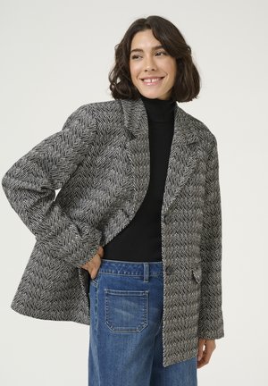 Mustavalkoinen herringbone-kuvioitu takki, jossa on viistetyt läpät, kaksi nappia ja teksturoitu kangas, käytetty mustan poolopaidan päällä.