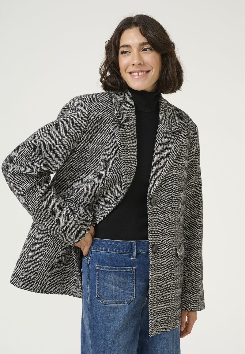 Black and white herringbone-patterned blazer with notched lapels, two buttons, and textured fabric, worn over a black turtleneck.