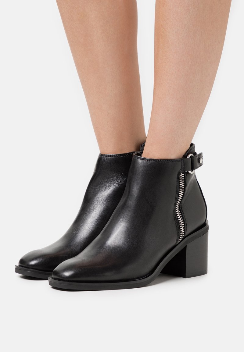 Bottes courtes en cuir noir avec talons carrés, fermeture éclair latérale et détail boucle, portées sur des jambes nues sur un fond blanc uni.