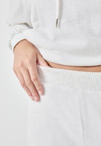 Witte, textuurstof van een sweatshirt met geribbelde zoom. Een hand rust op de bijpassende tailleband, waarmee het zachte materiaal en het ontwerp worden getoond.