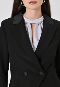 Blazer nero con revers in satin, design doppiopetto e due bottoni neri. Sotto, una blusa grigio chiaro con apertura a chiave.