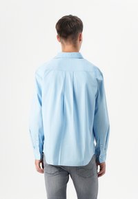 Junger Mann steht mit dem Rücken zur Kamera, trägt ein hellblaues Hemd und graue Jeans, vor einem einfarbigen weißen Hintergrund.