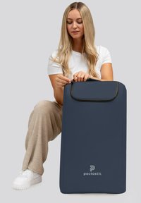 Donna con lunghi capelli biondi che indossa una camicia bianca e pantaloni beige mentre apre una grande borsa navy blue Pactastic su sfondo grigio.