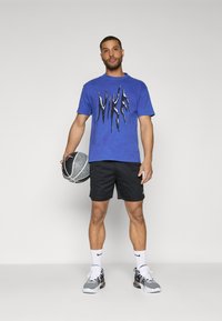 Camiseta de algodón azul con un diseño gráfico negro, pantalones cortos negros y zapatillas grises. Sosteniendo un balón de baloncesto con un patrón, de pie frente a un fondo neutro.