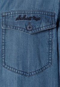 Denimhemd mit einer Brusttasche, hellblauer Farbe, kontrastierenden Nähten und einem dunklen Knopf mit "Schott NYC" oben auf der Tasche bestickt.