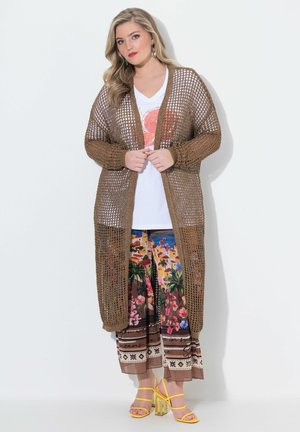 Femme portant un long cardigan marron en maille ajourée, un t-shirt blanc avec un motif graphique, un pantalon large à fleurs colorées et des talons jaunes à brides sur fond blanc.
