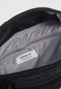 Černý batoh uvnitř s šedým podšívkou, obsahuje zipovou kapsu a štítek se zobrazeným logem Adidas a informacemi o značce.
