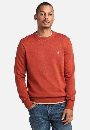 Maglione rosso a maglia con colletto rotondo e polsini a coste, con un piccolo logo sul petto. Indossato sopra una t-shirt chiara e jeans di denim.