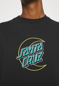 Black T-shirt featuring an embroidered "Santa Cruz" logo in turquoise and white, outlined with a yellow crescent shape. Soft fabric texture.