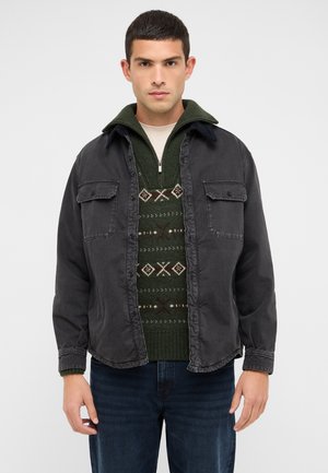 Dunkelgraue Button-Down-Jacke mit zwei Brusttaschen, über einem grün gemusterten Pullover getragen, kombiniert mit dunklen Jeans. Strukturierte, lässige Designs.