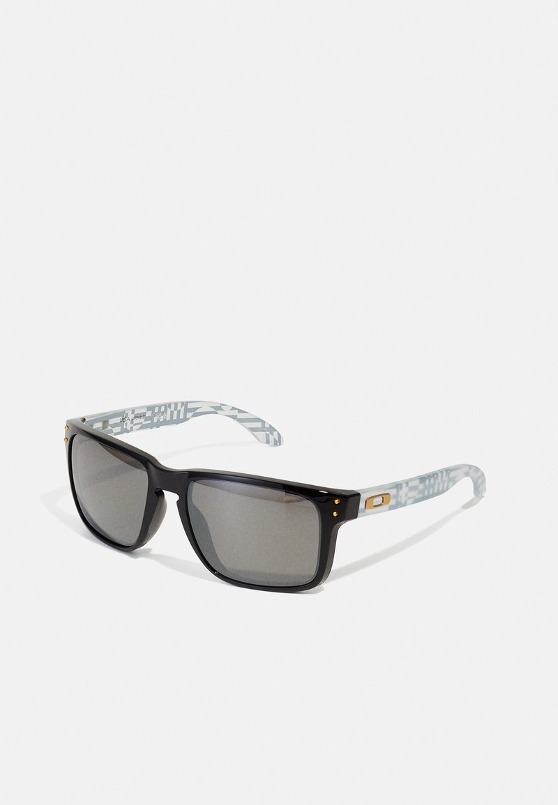 Occhiali da sole neri con bracci a motivo grigio, lenti scure e dettagli in oro sulle tempie. Montatura resistente con un design squadrato.