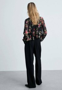 Blusa nera floreale con orlo arricciato, materiale trasparente; abbinata a pantaloni neri a gamba dritta. Modella vista da dietro.