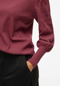 Pull en tricot bordeaux avec des manches texturées et bouffantes et des poignets ajustés, associé à un pantalon noir. Il présente une finition lisse et une coupe décontractée.