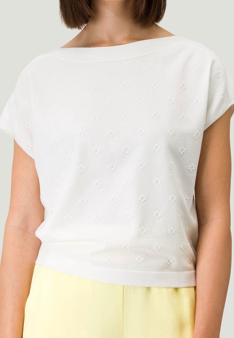 Hvid kortærmet top med bådudskæring, prydet med teksturerede blomster mønstre. Blødt strikket materiale med en afslappet pasform og minimalistisk design.