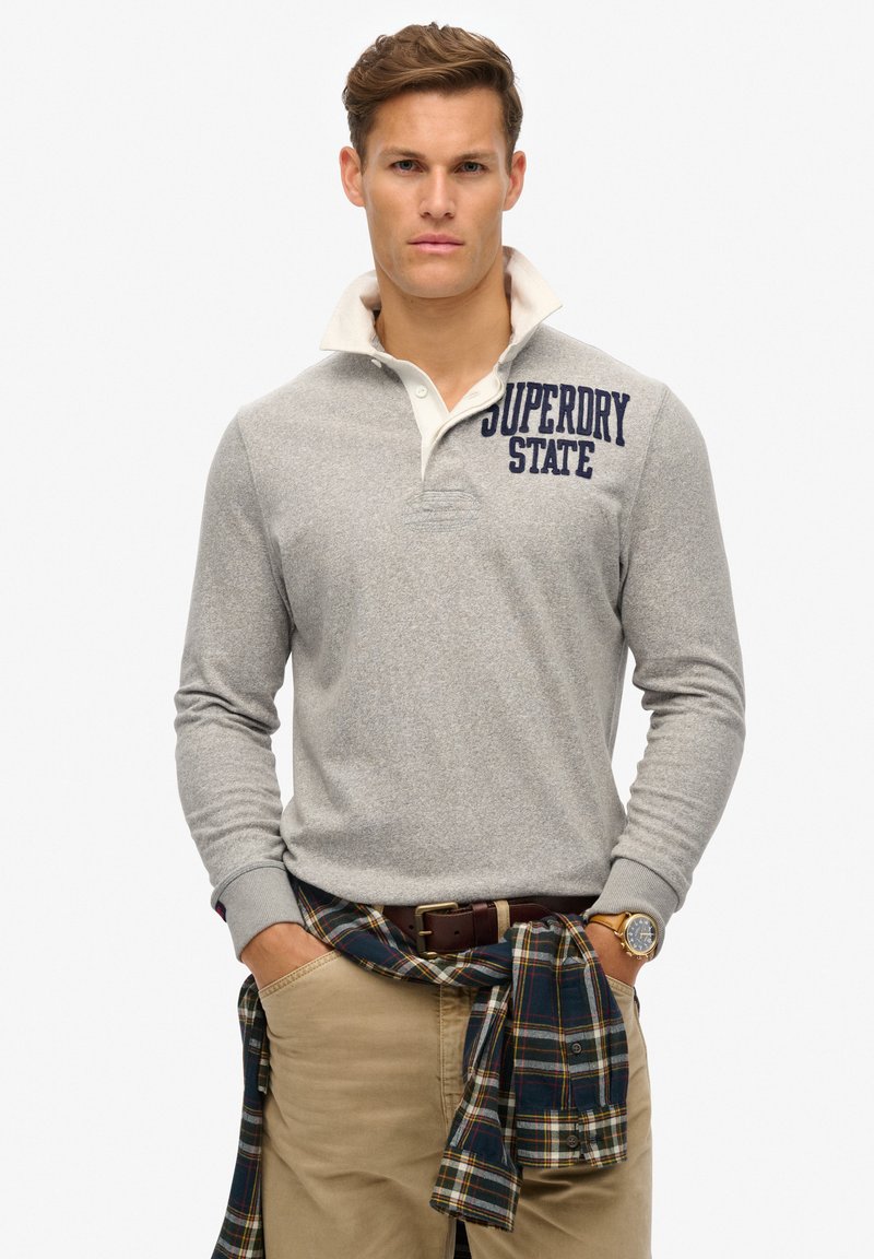 Polo gris de manga larga con cuello blanco, lleva el logo "Superdry State" en azul marino. Pantalones beige con una camisa a cuadros atada a la cintura, cinturón marrón y reloj.