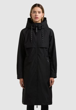 Mujer con un impermeable largo negro con capucha, cierre frontal y cordón en la capucha, de pie contra un fondo liso.