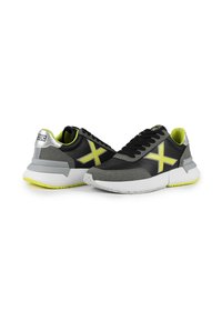 Baskets de sport noires et grises avec des accents jaunes vifs. Comprend une tige texturée, des panneaux en mesh et une semelle blanche avec un design façon éclaboussures.