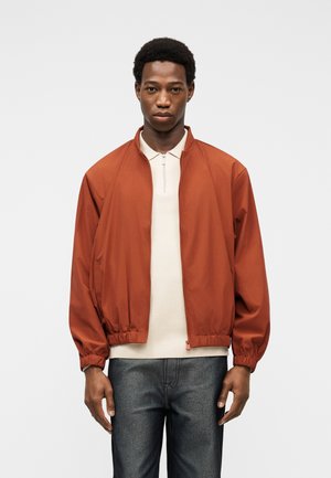 Homme portant une veste zippée orange rouille par-dessus une chemise beige à col et un jean bleu foncé, debout devant un fond clair uni.
