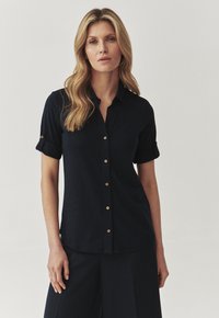 Camisa de botones color azul marino con mangas cortas, puños enrollados y detalles de herrajes dorados. Fabricada en una tela relajada y texturizada.