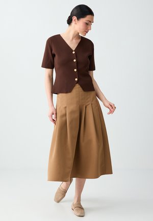 Woman wearing a brown short-sleeve buttoned top, tan pleated midi skirt, and beige slip-on shoes, standing against a plain light background.
