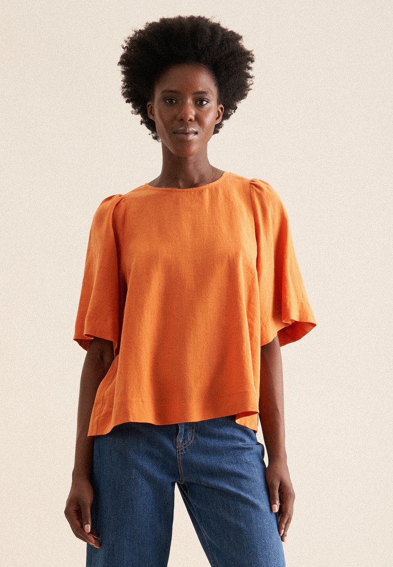 Top in lino arancione a vestibilità ampia con maniche corte a campana e scollatura rotonda, abbinato a jeans in denim blu. Tessuto testurizzato.