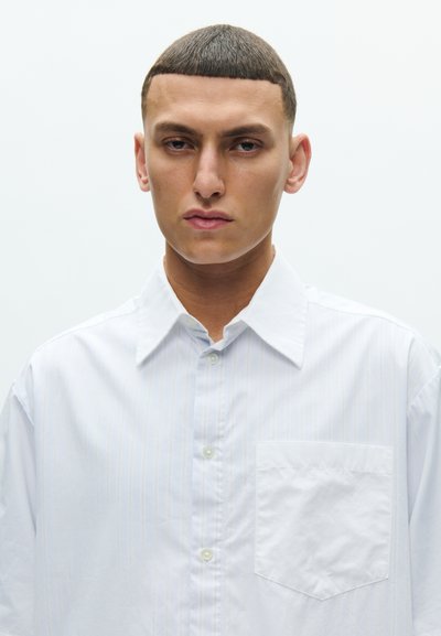 Mladý muž s krátkymi tmavými vlasmi, oblečený v bielej košeli na gombíky, smerujúci vpred na jednoduchom svetlom pozadí.