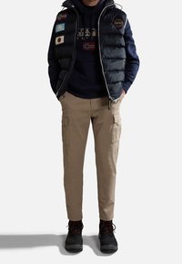 Granatowa, ocieplana kamizelka z naszywkami w formie flag, noszona na ciemnej bluzie z kapturem z logo. Do tego jasnobrązowe spodnie cargo i czarne buty z czerwonymi akcentami dopełniają strój.