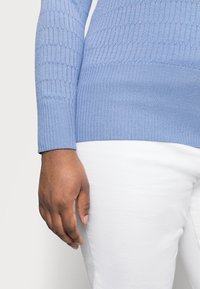 Hellblauer Strickpullover mit strukturierter Wabenmusterung an den Ärmeln. Gerippte Bündchen an Ärmeln und Saum. Kombiniert mit weißen Hosen, auf denen eine Hand ruht.