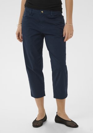 Žena, ktorá má na sebe tmavomodré skrátené nohavice, čierny top a čierne tkané nazúvacie topánky, stojaca na jednoduchom svetlom pozadí.