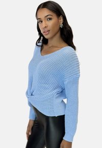 Hellblaues Strickpullover in lockerer Passform, mit einem Twist-Detail an der Taille und langen Ärmeln. Kombiniert mit schwarzen Leggings.