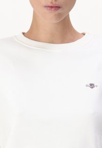 Femme portant un sweat-shirt blanc col rond avec un petit logo GANT brodé représentant un bouclier et des étoiles sur le côté gauche de la poitrine.